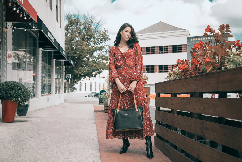 Fashionable Gifts For Working Moms - A woman in a red dress stands next to a flower bed with her black and hazelnut Emily Tote Bag. 